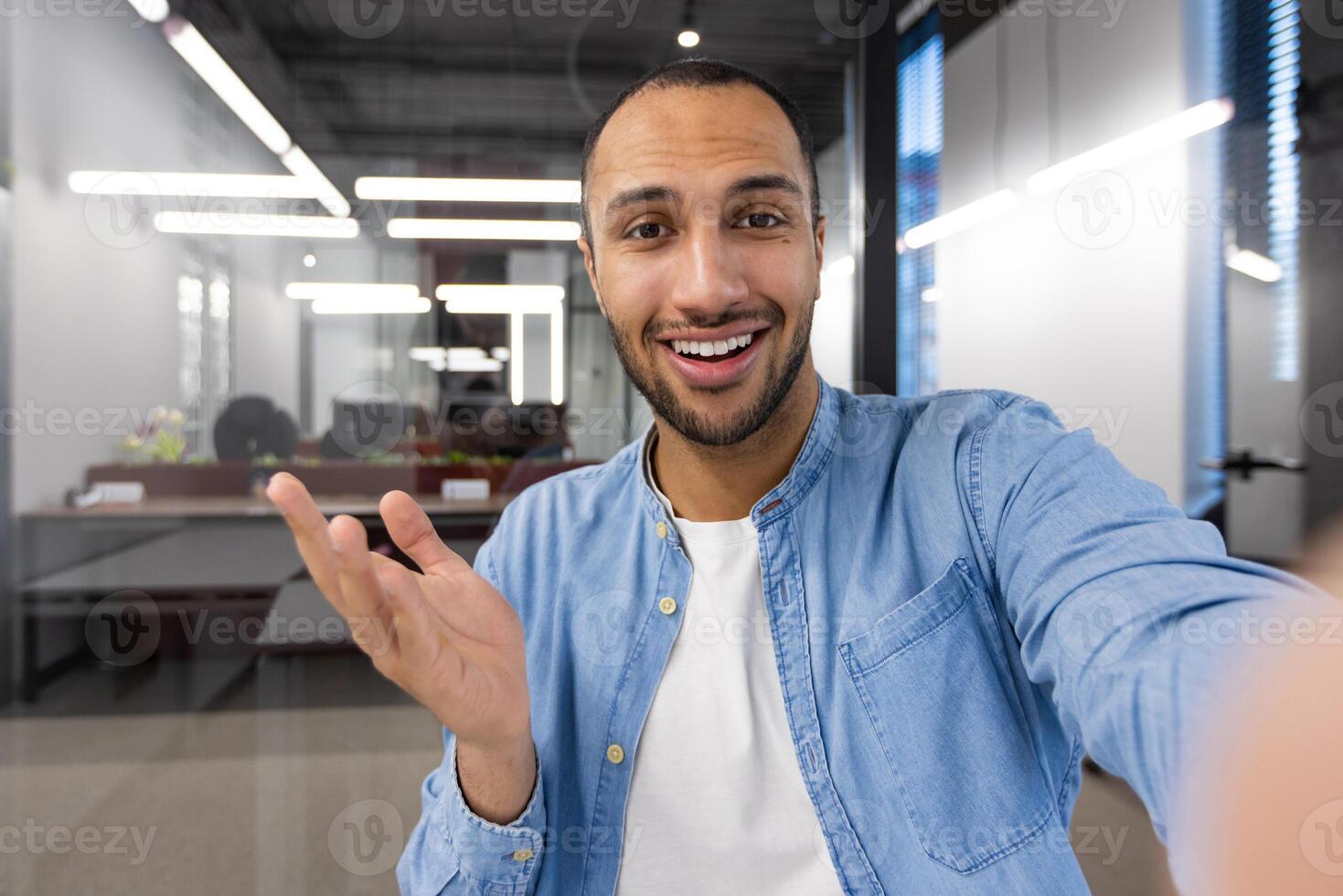 A cheerful young man capturing a selfie with a smartphone in a well-lit contemporary office setting, expressing positivity and confidence. photo