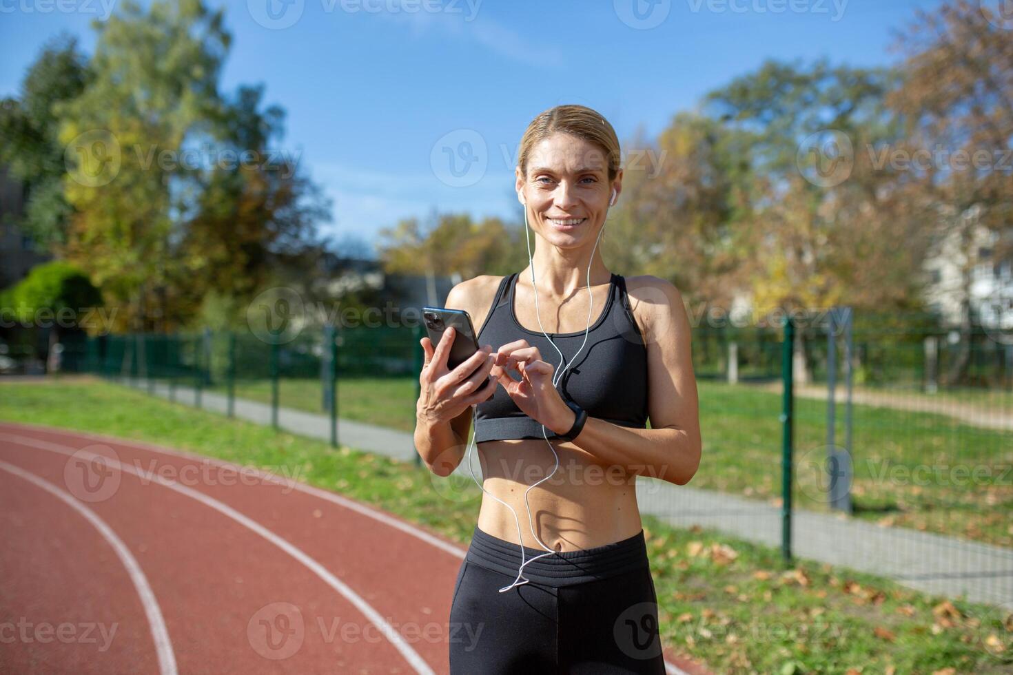 Fit woman in sportswear on a running track choosing songs on her phone, with earphones, ready for a workout. photo