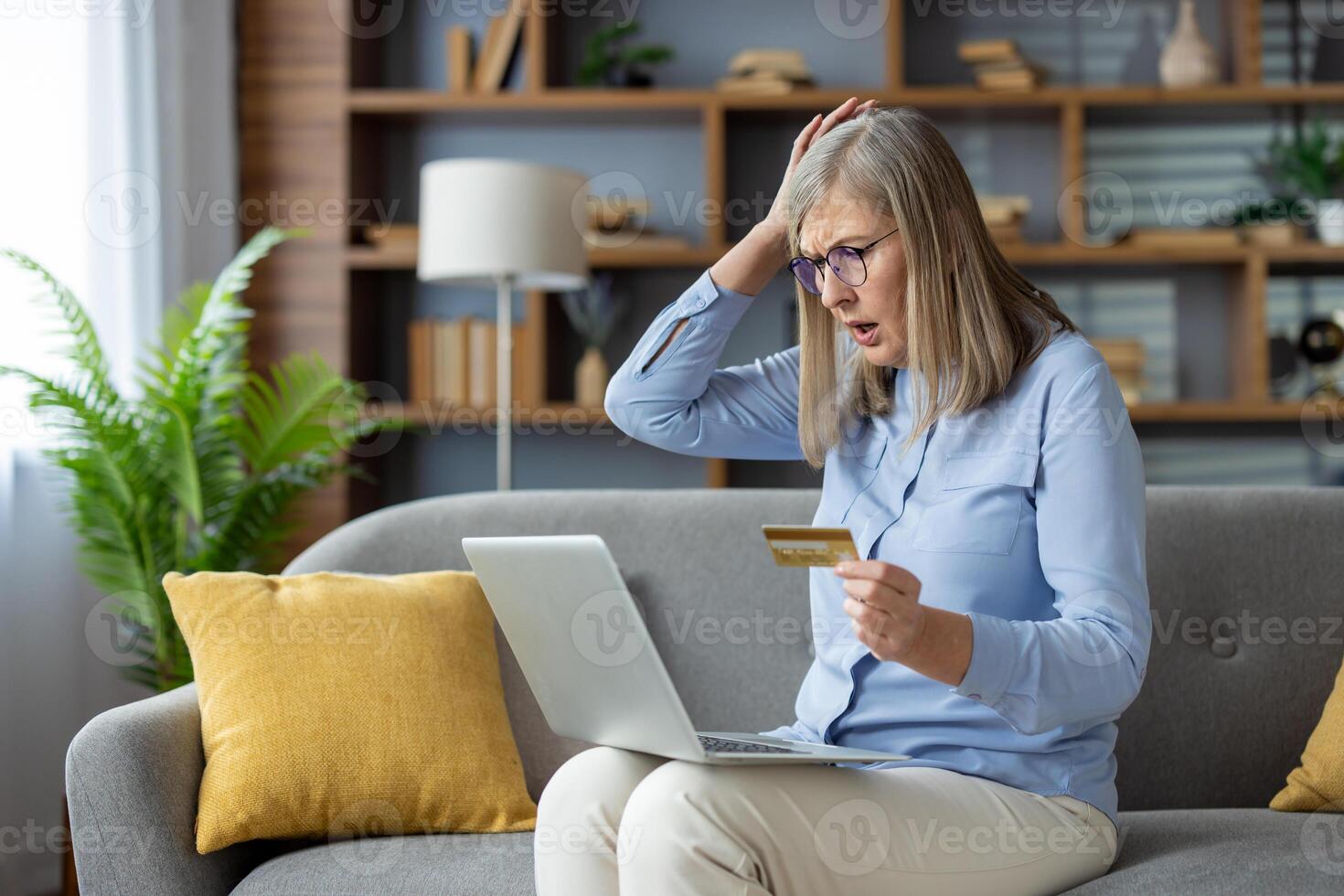 A mature woman looking surprised and anxious while holding a credit card and using a laptop on her sofa at home. photo