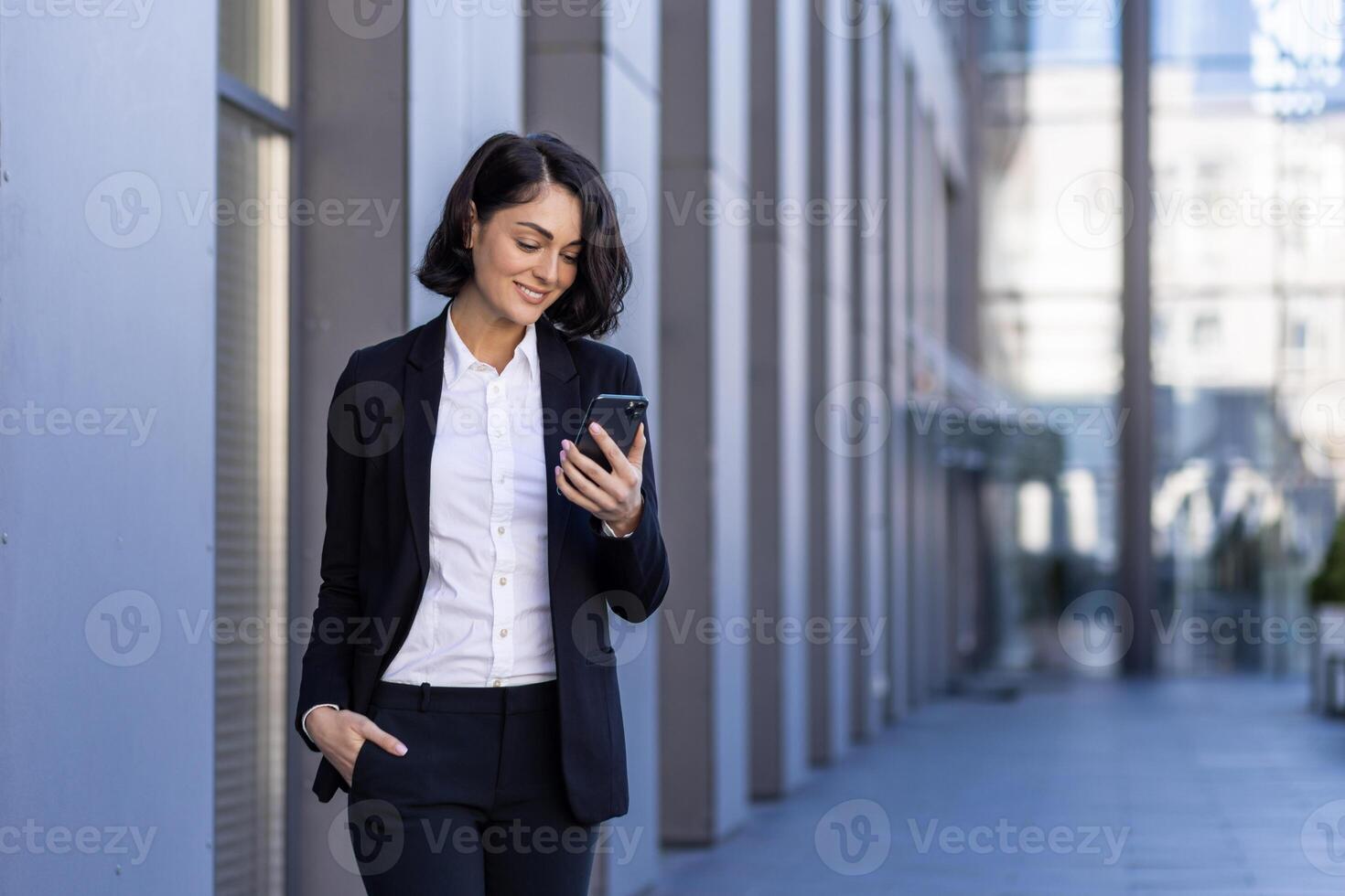A young beautiful business woman is walking outside the office building, a woman in business clothes is smiling, using an application on the phone, browsing the Internet, typing a message. photo