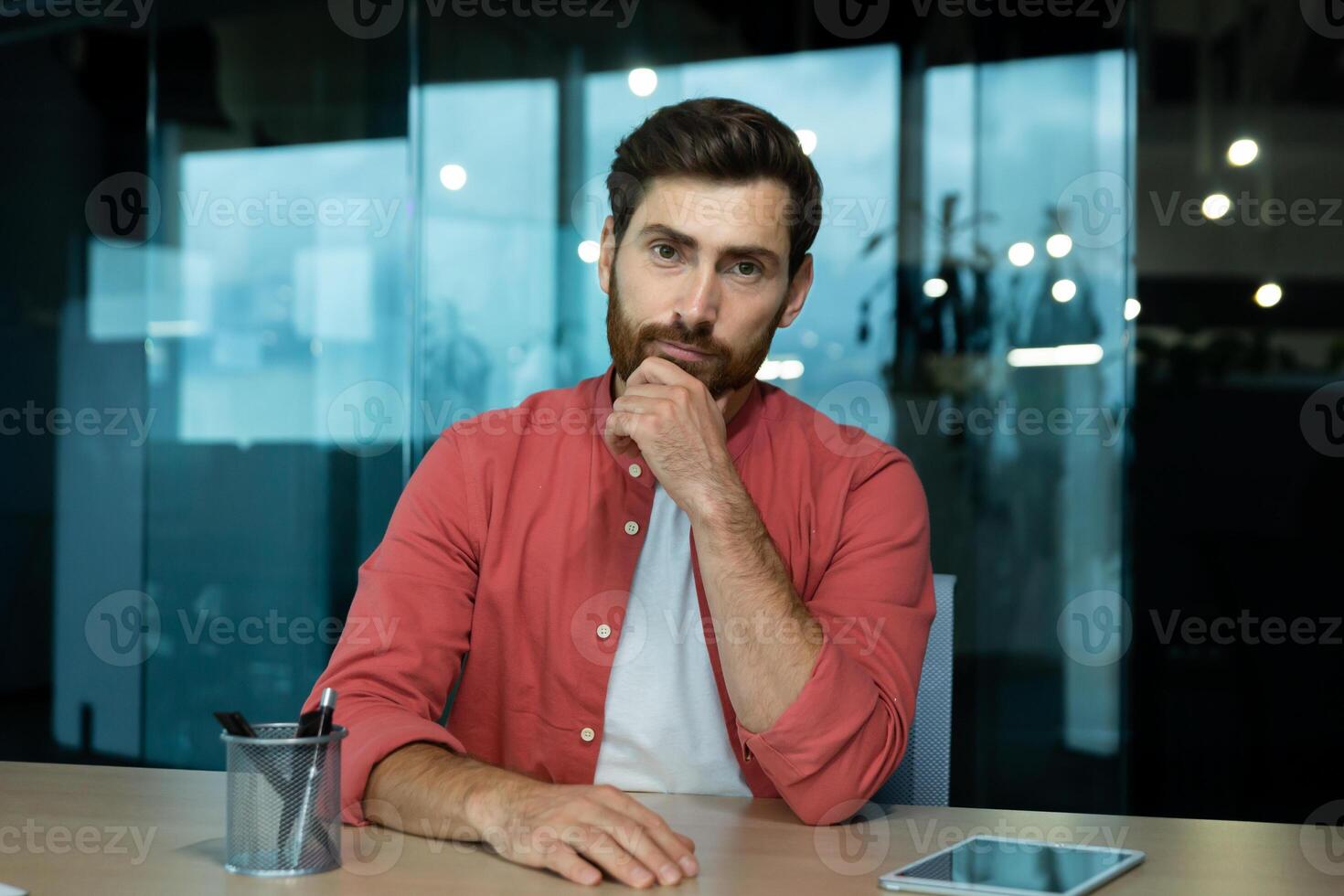 Online chat, remote business meeting, businessman in red shirt looking seriously thinking and focused into camera, listening to interlocutor, webcam view, inside office. photo