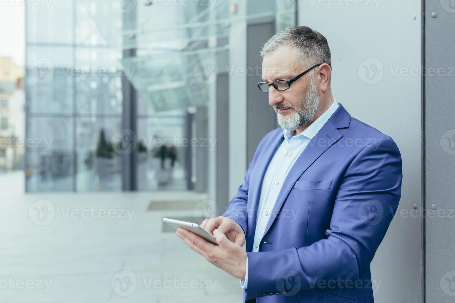 Senior handsome businessman man in glasses and a suit stands on the street near the office center and waits for customers, employees, holds the phone in his hands, dials a number, messages. photo