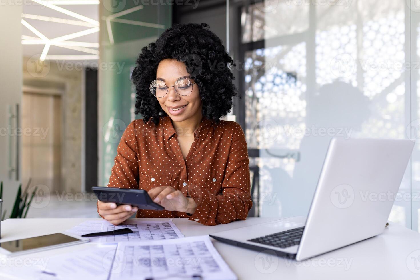 Young beautiful businesswoman, paper work inside office, female employee uses calculator at work, calculates and checks reports, accountant financier with laptop, reviews documents, uses laptop. photo