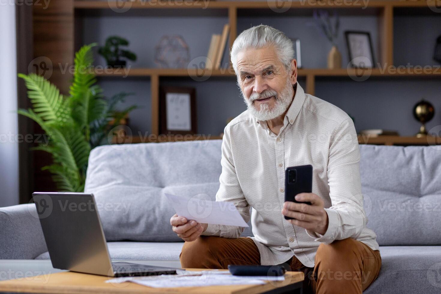 Elderly bearded man sitting on a couch, holding documents and smartphone while working with a laptop. Comfortable home office setup in a cozy living room. photo