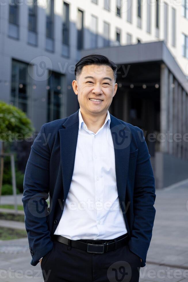 Portrait of an Asian businessman outdoors, featuring a confident smile, dressed in formal wear, with a modern building background. photo