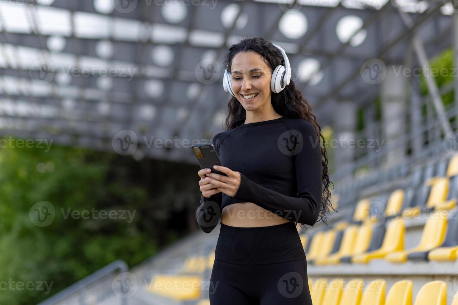 un joven mujer en atlético vestir con auriculares participación un teléfono inteligente mientras en pie en un estadio. concepto de aptitud y moderno estilo de vida. foto