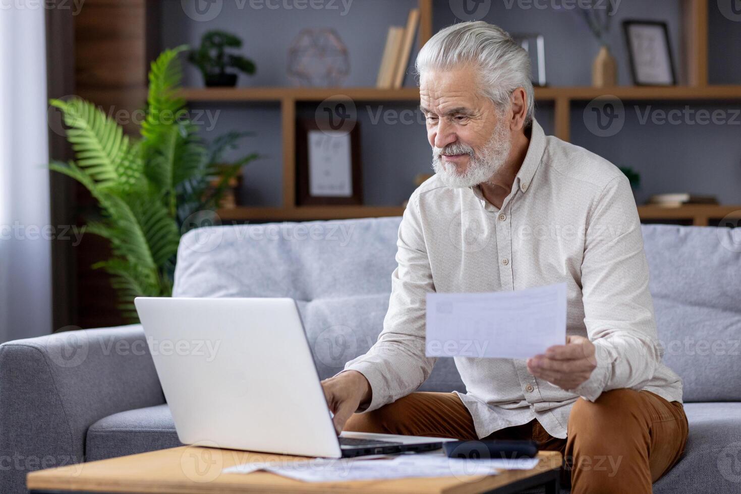mayor de pelo blanco hombre en un casual camisa mira a papeleo mientras sentado en un sofá, utilizando un ordenador portátil en un bien amueblado vivo habitación. foto