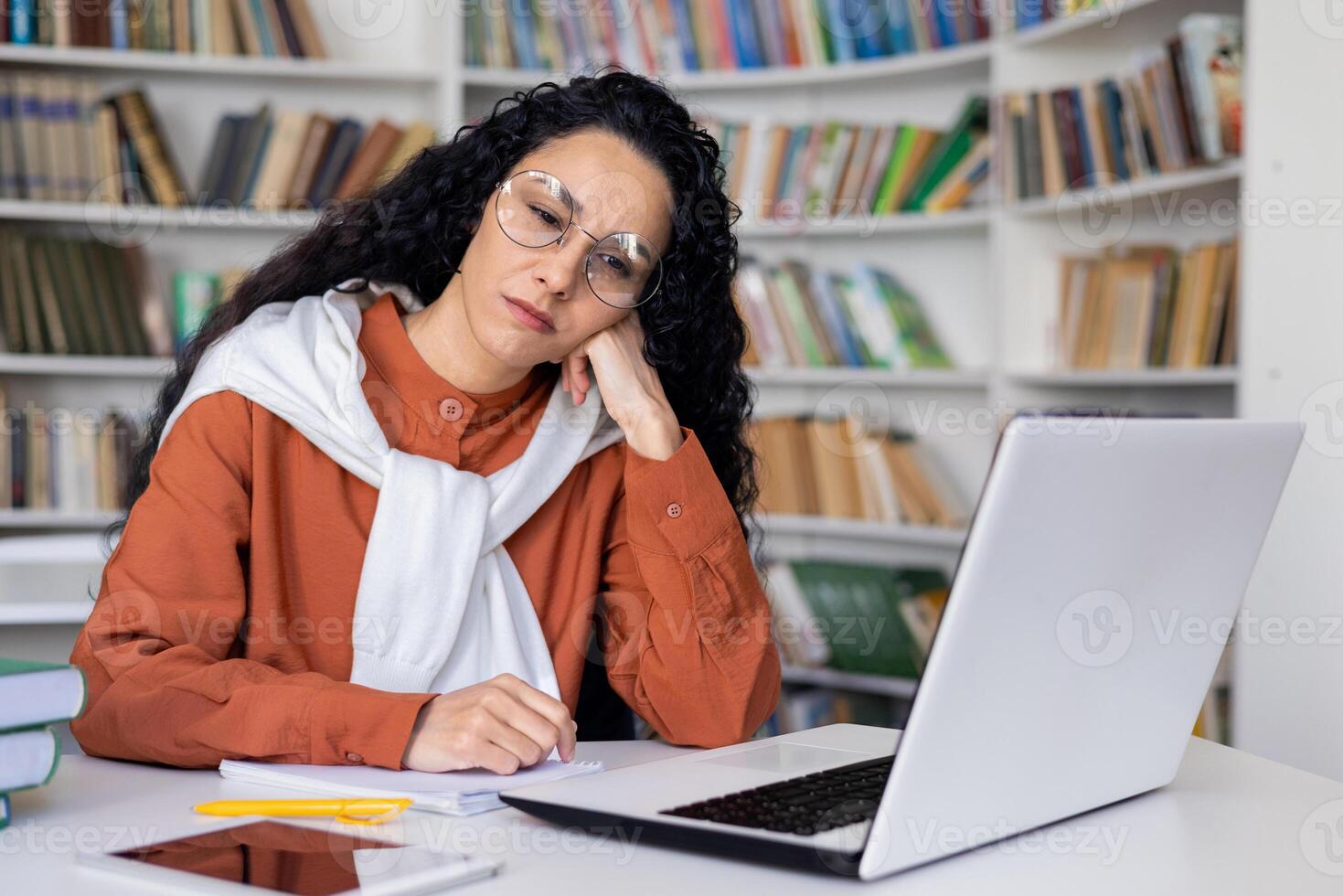 Bored caucasian female sitting at desk with modern laptop and working with paper materials with books in front of them. Upset woman with glasses feeling tired from monotone work process. photo