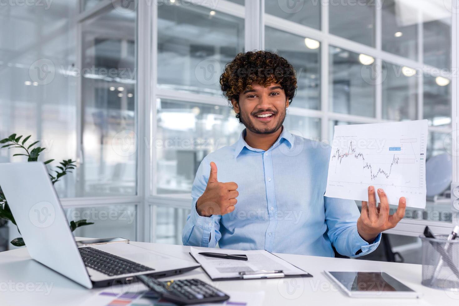 Portrait of young successful financier paperwork inside office, man smiling and looking at camera shows graph with positive dynamics of economic profits, businessman at work with laptop. photo