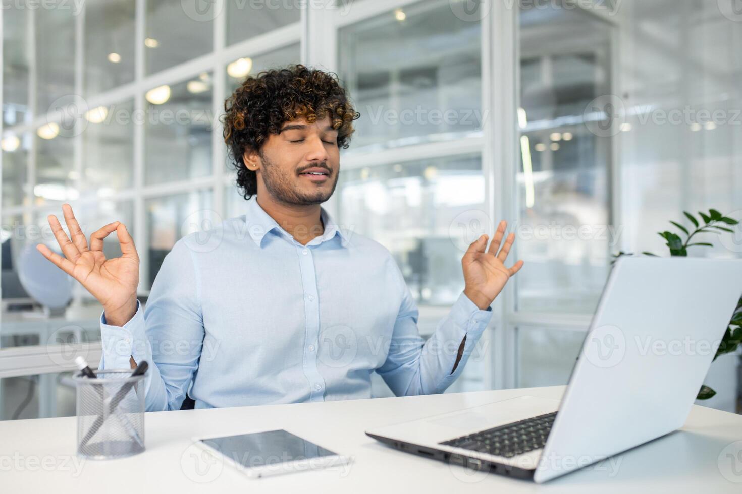 Young businessman meditating at his workplace in a busy office. He uses mindfulness to reduce stress, sitting with eyes closed and hand in a mudra position. photo