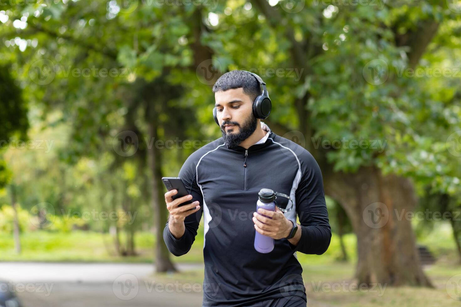 un ajuste hombre en ropa de deporte toma un descanso en un lozano parque a utilizar su teléfono inteligente y bebida agua, encarnando un activo, sano estilo de vida. foto