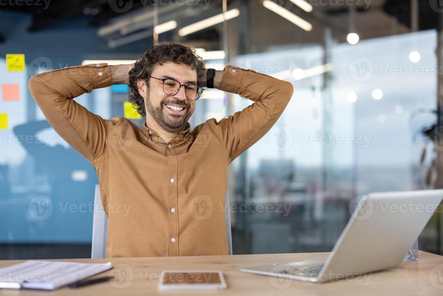 Well done businessman successfully completed work project inside office at workplace, man with hands behind head resting happily smiling , looking at laptop screen satisfied with achievement results photo