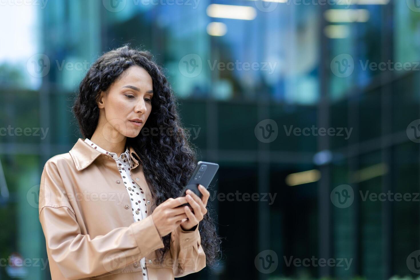 grave negocio mujer con teléfono en manos, latín americano mujer pensando mecanografía mensaje y leyendo noticias, jefe utilizando aplicación en teléfono inteligente mientras caminando fuera de oficina edificio. foto