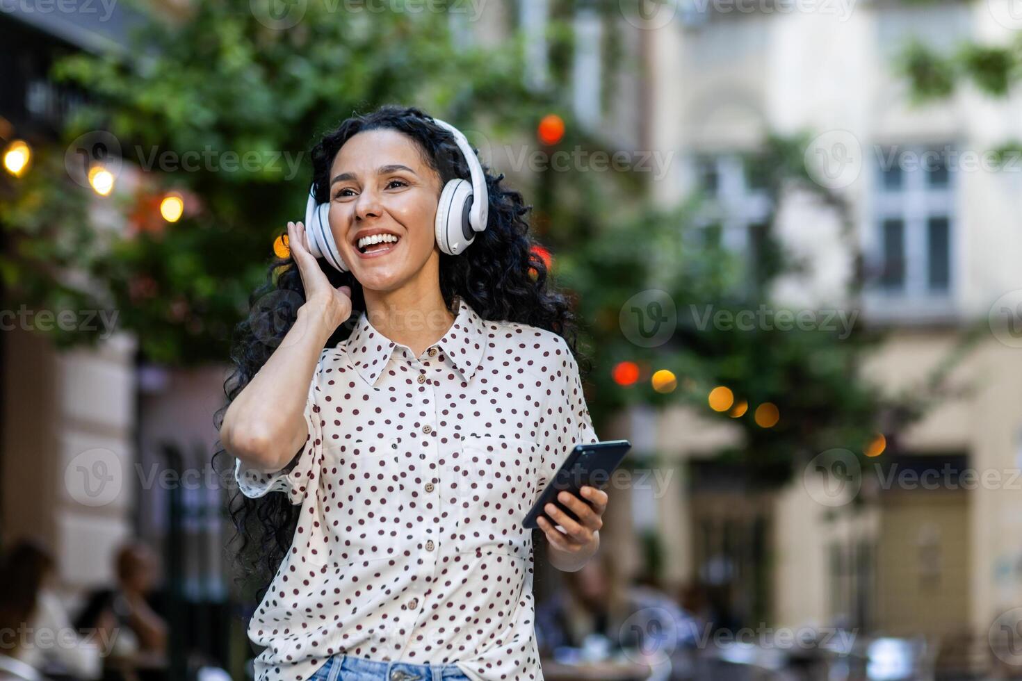 Beautiful young Latin American woman walks in the evening city on a trip, woman with headphones listens to music and dances and sings, uses an online radio application on her phone photo
