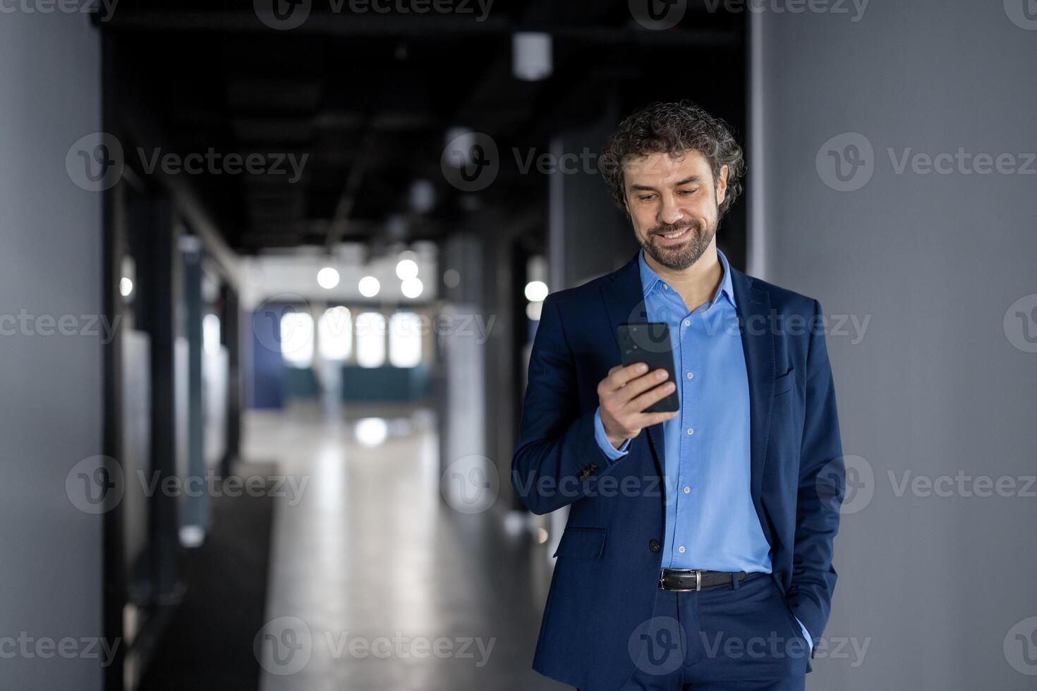 maduro experimentado exitoso empresario en negocio traje caminando dentro oficina edificio, hombre caminando abajo corredor con teléfono en manos, jefe utilizando aplicación teléfono inteligente, leyendo y sonriente contentamente foto