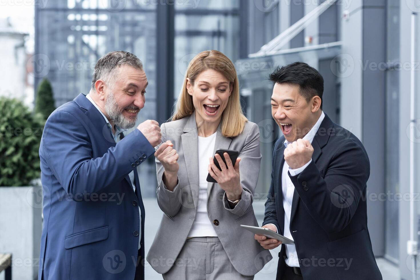 group of colleagues employees read good news looking at smartphone. Happy team rejoices about successful investment while using, browsing mobile phone. Standing outside near office building. photo