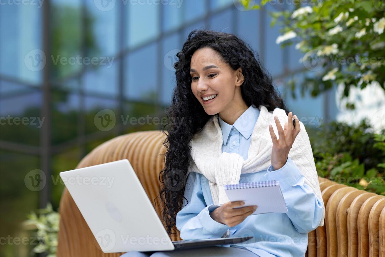 Young beautiful woman sits on a bench outside an office building, uses a laptop to watch a webinar course , a student writes data in a notebook, call with a teacher, distance learning. photo