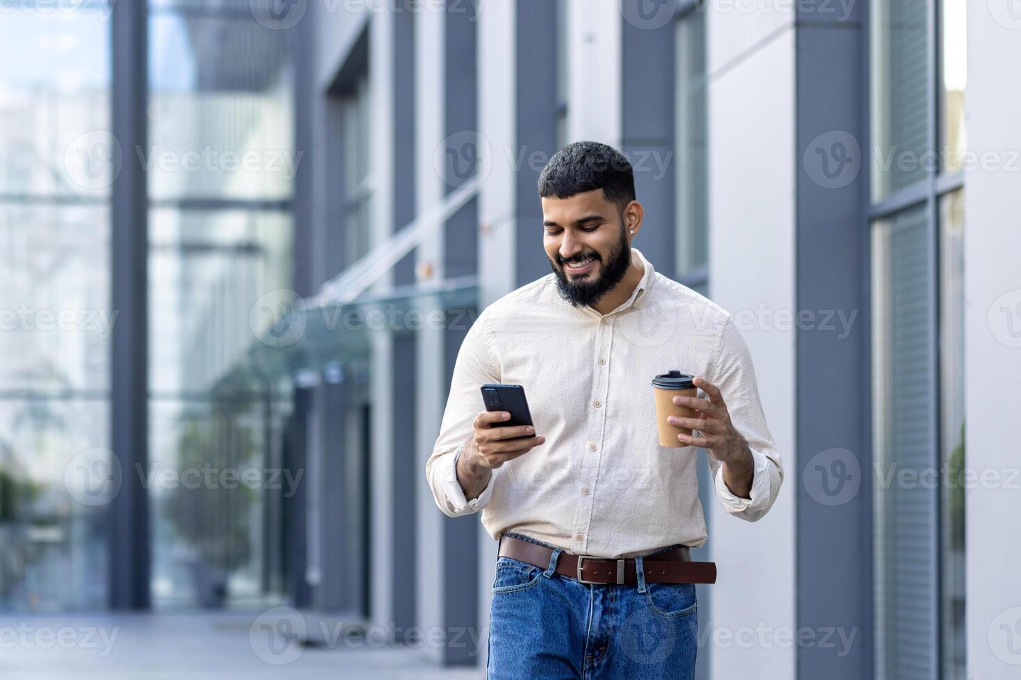 un alegre barbado joven adulto es absorto en su teléfono inteligente mientras por casualidad participación un para llevar café taza. él es vagante en frente de un moderno vaso edificio, ejemplificando un relajado aún dinámica urbano estilo de vida. foto