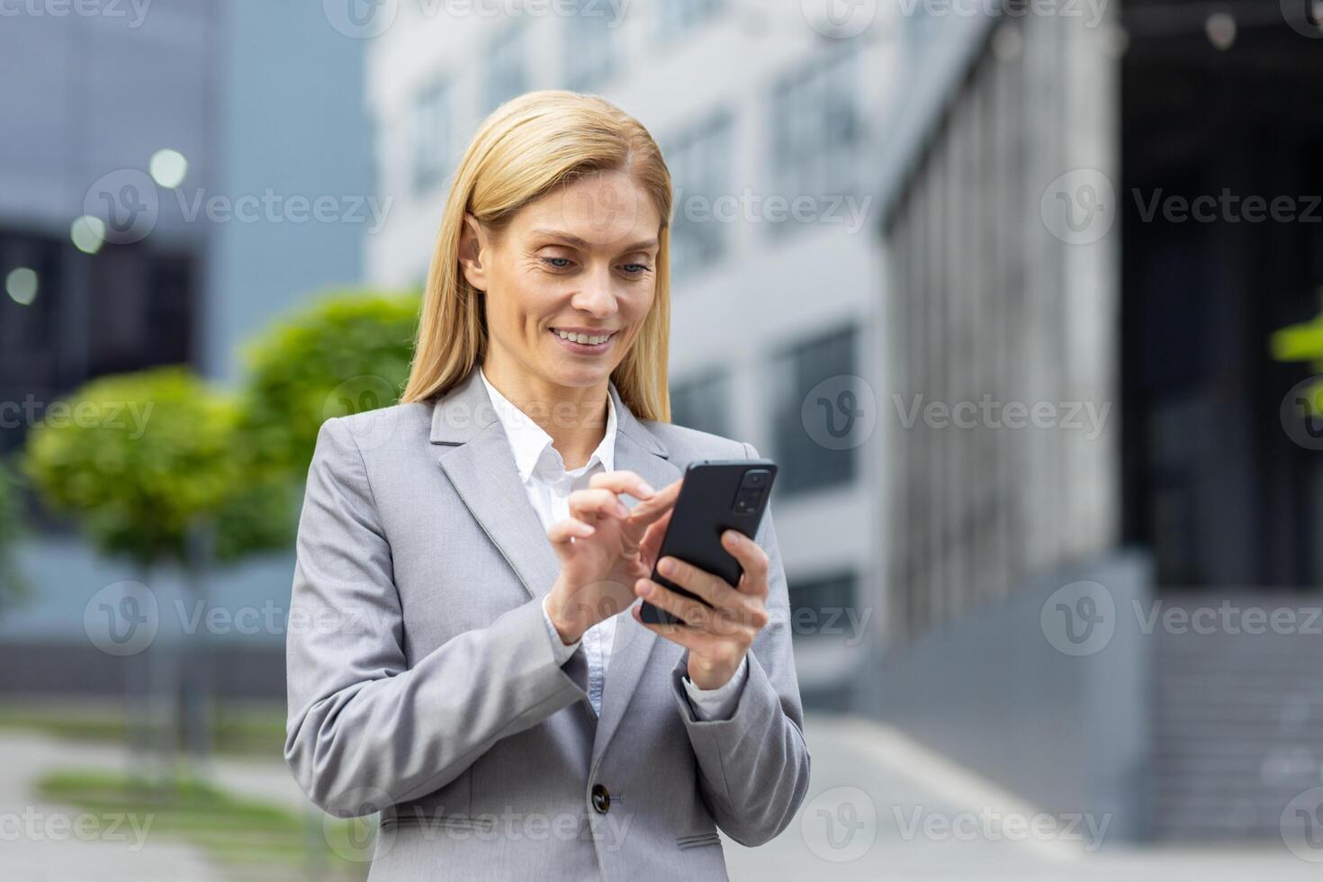 Successful woman boss walks outside office building outdoors, in business suit, businesswoman uses app on phone, smiles contentedly reads internet pages, browses news, writes text message, call. photo