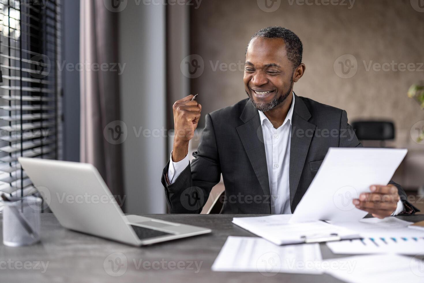 alegre masculino empresario expresando felicidad en oficina configuración, puño bomba gesto con papeleo y ordenador portátil. foto