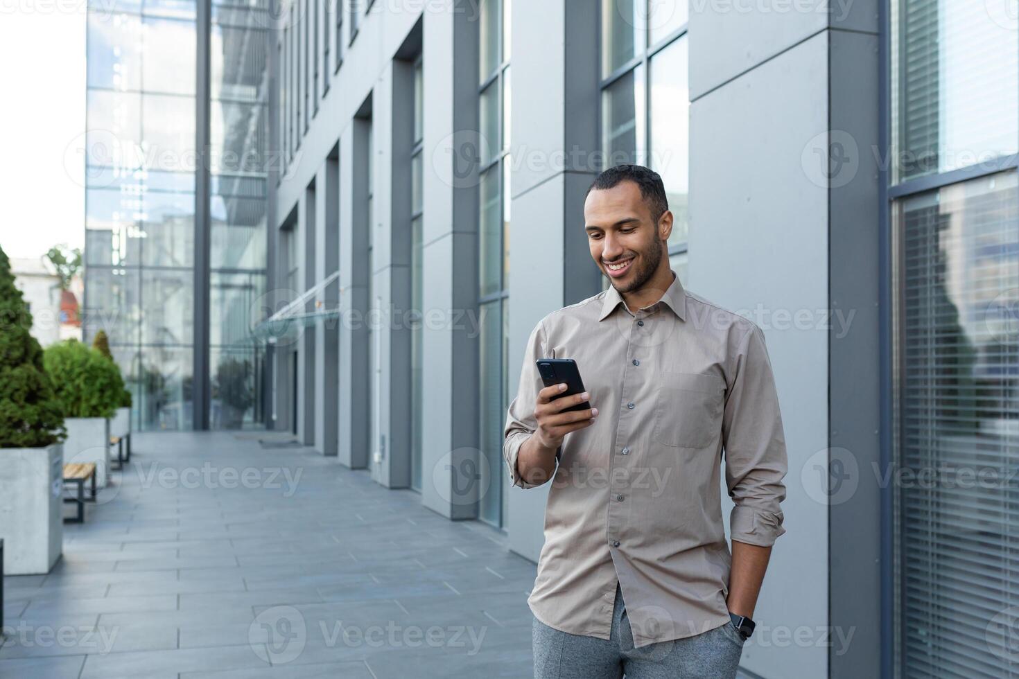 latín americano empresario en casual camisa caminando cerca oficina edificio desde afuera, hombre sonriente y utilizando teléfono, persona de libre dedicación mecanografía mensaje y hojeada Internet paginas foto