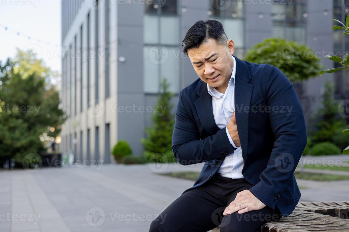 joven asiático masculino empresario sentado doblado fuera de oficina edificio en traje, participación mano a cofre y sensación grave dolor en corazón. foto