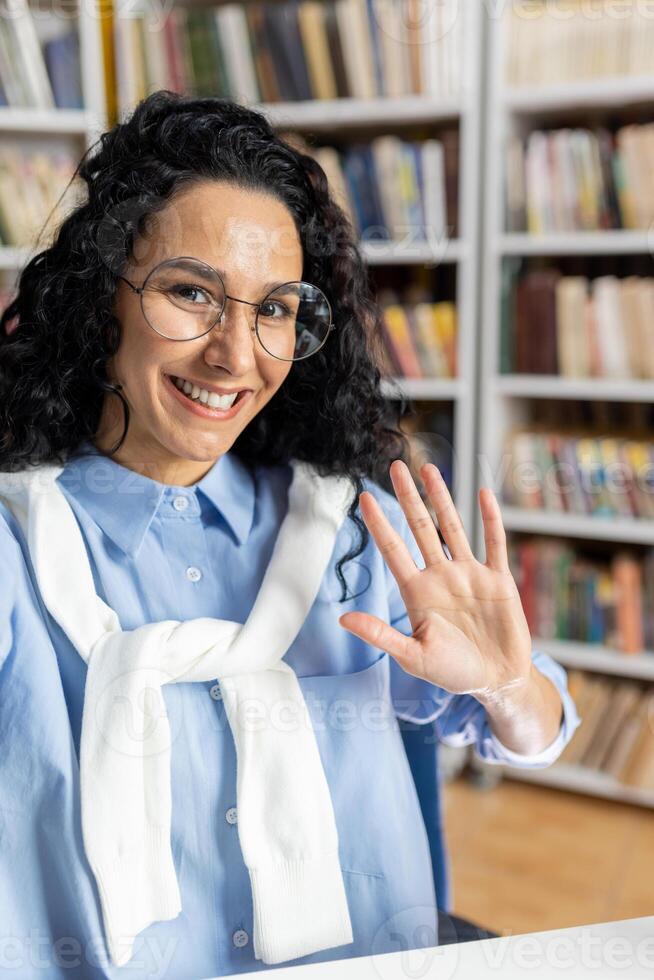 A cheerful woman with curly hair, wearing glasses and a casual outfit, happily waves in a well-stocked library, surrounded by books. photo