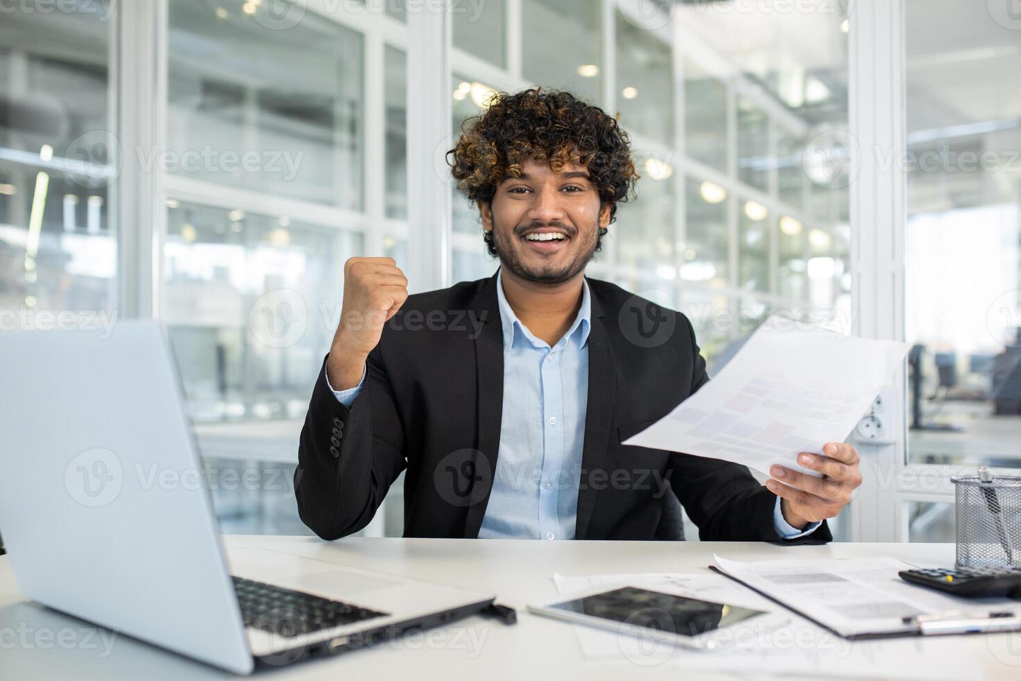 un alegre joven hombre en un negocio traje celebra un exitoso proyecto o logro mientras sentado a su escritorio en un moderno oficina ambiente, demostración victoria puño y sonriente con documentos en mano. foto