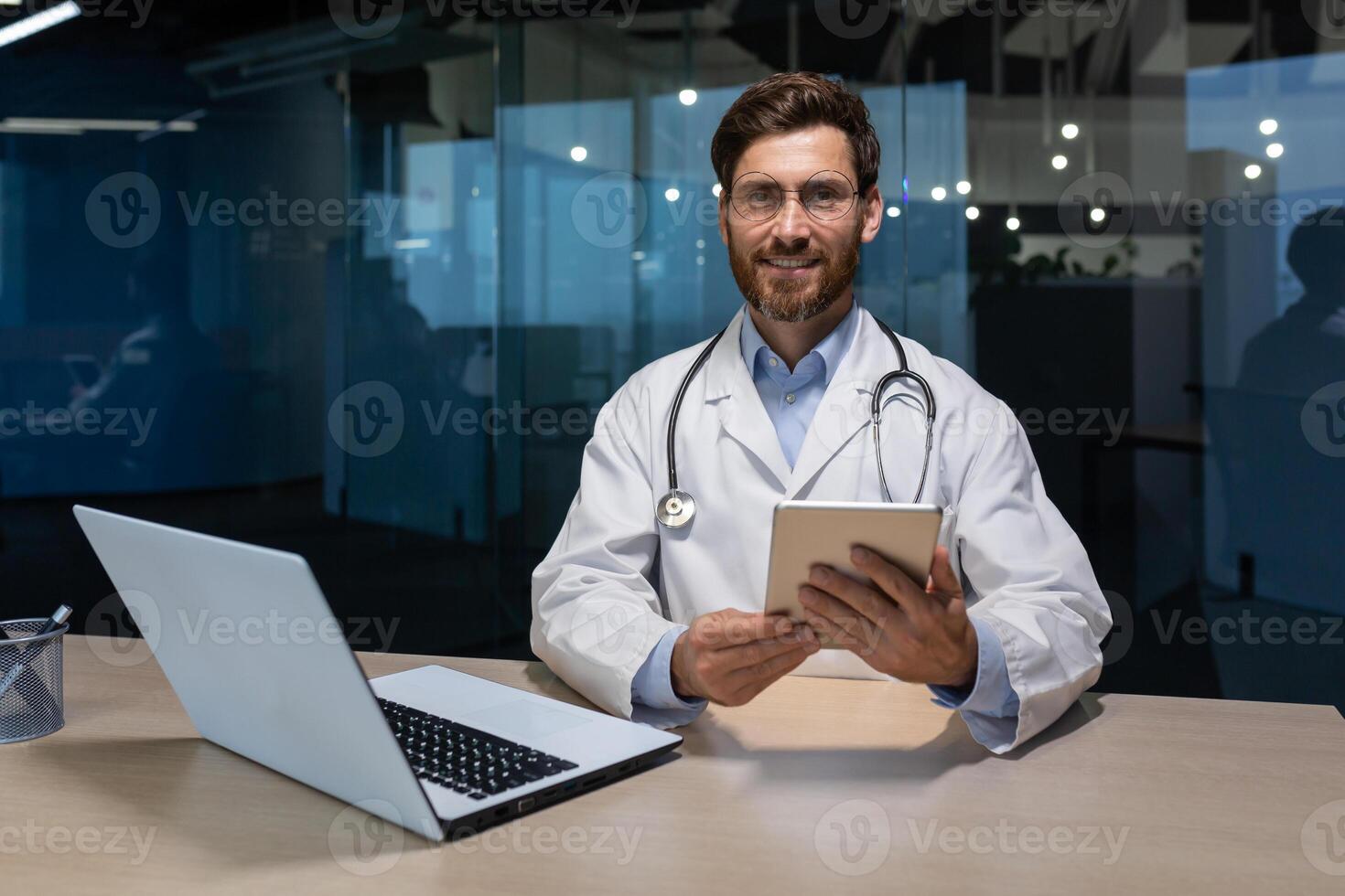 Portrait of a young handsome male doctor who sits and works in an office in a hospital at a table with a laptop. Holds a tablet in his hands, looks at the camera, smiles. photo