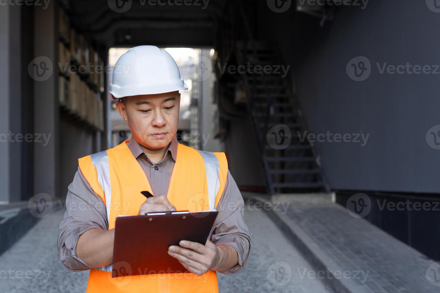 Concentrated and serious young Asian male architect, builder. the engineer stands outside in a hard hat and vest and makes notes in documents, conducts inspections, draws up a project plan. photo