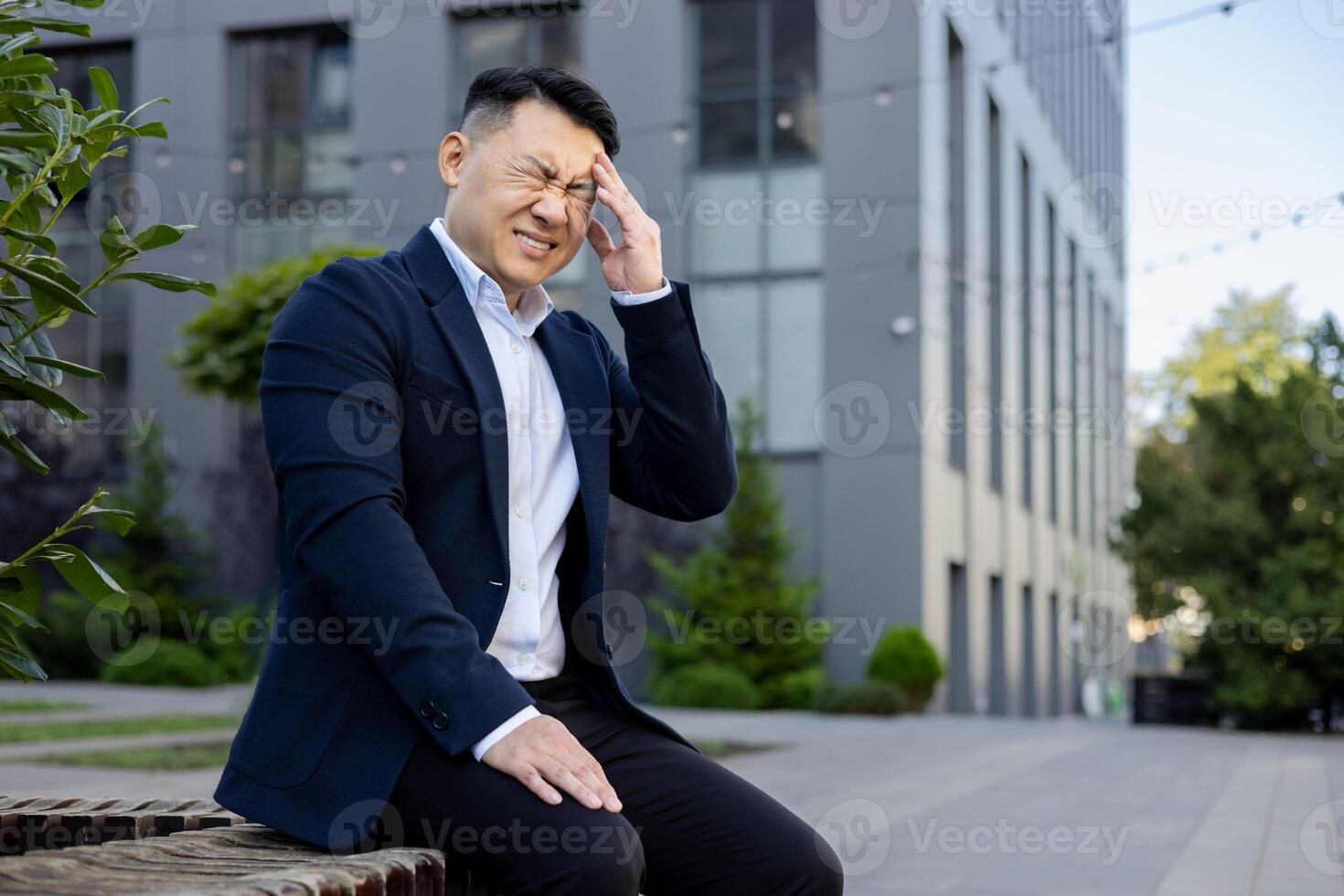 A professional male in a suit looks troubled, with one hand on his head, expressing headache or stress outdoors. photo