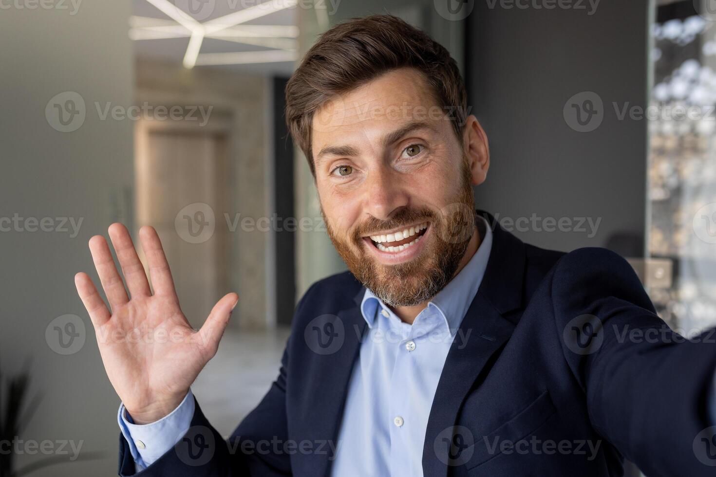 Close-up portrait of a smiling and successful young businessman in a suit standing in the office, waving and greeting with his hand on the camera phone, talking on call. photo