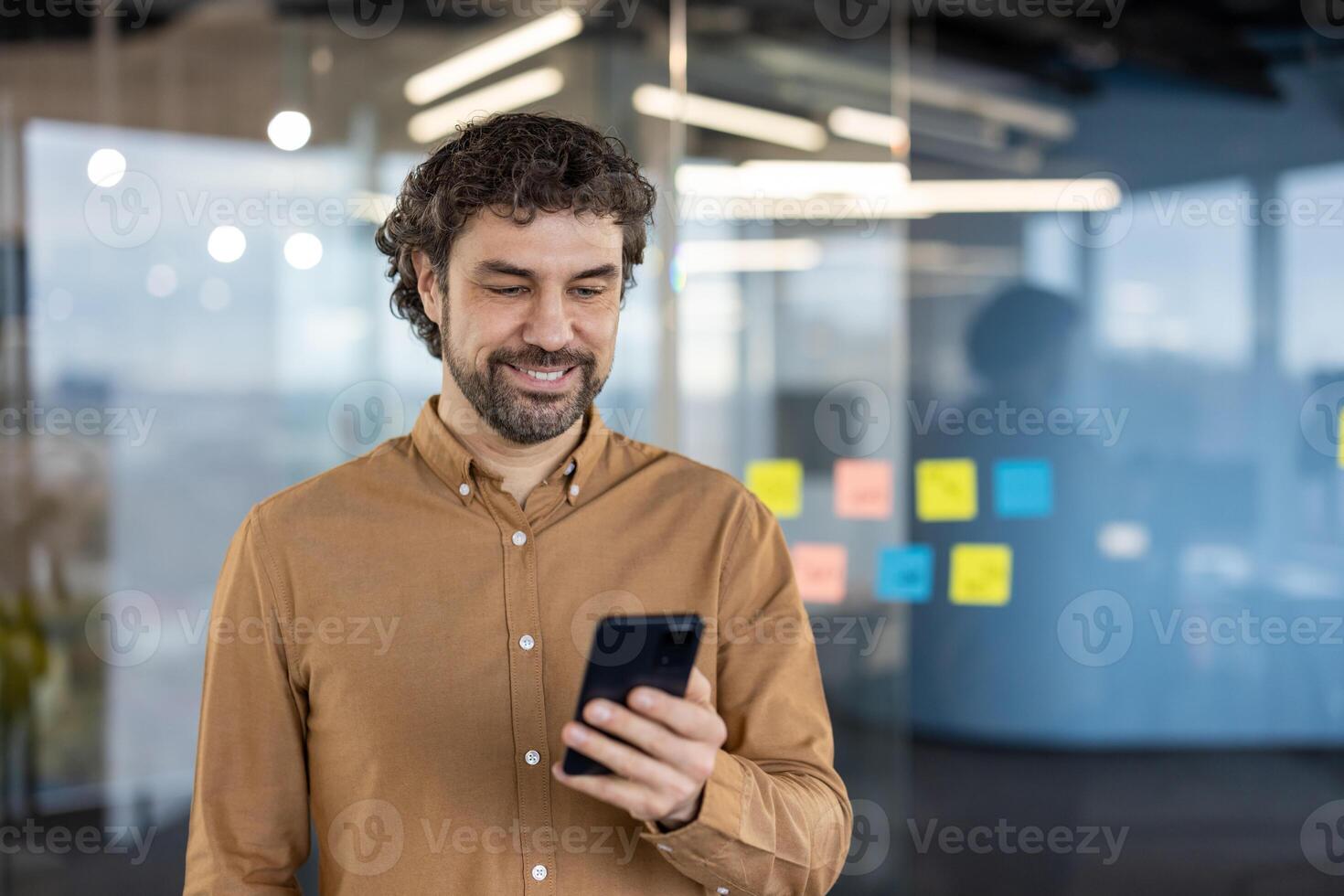 Carefree guy with stylish hairstyle using black cell phone while standing alone in working space. Relaxed man watching interesting in social media while using wireless internet connection. photo