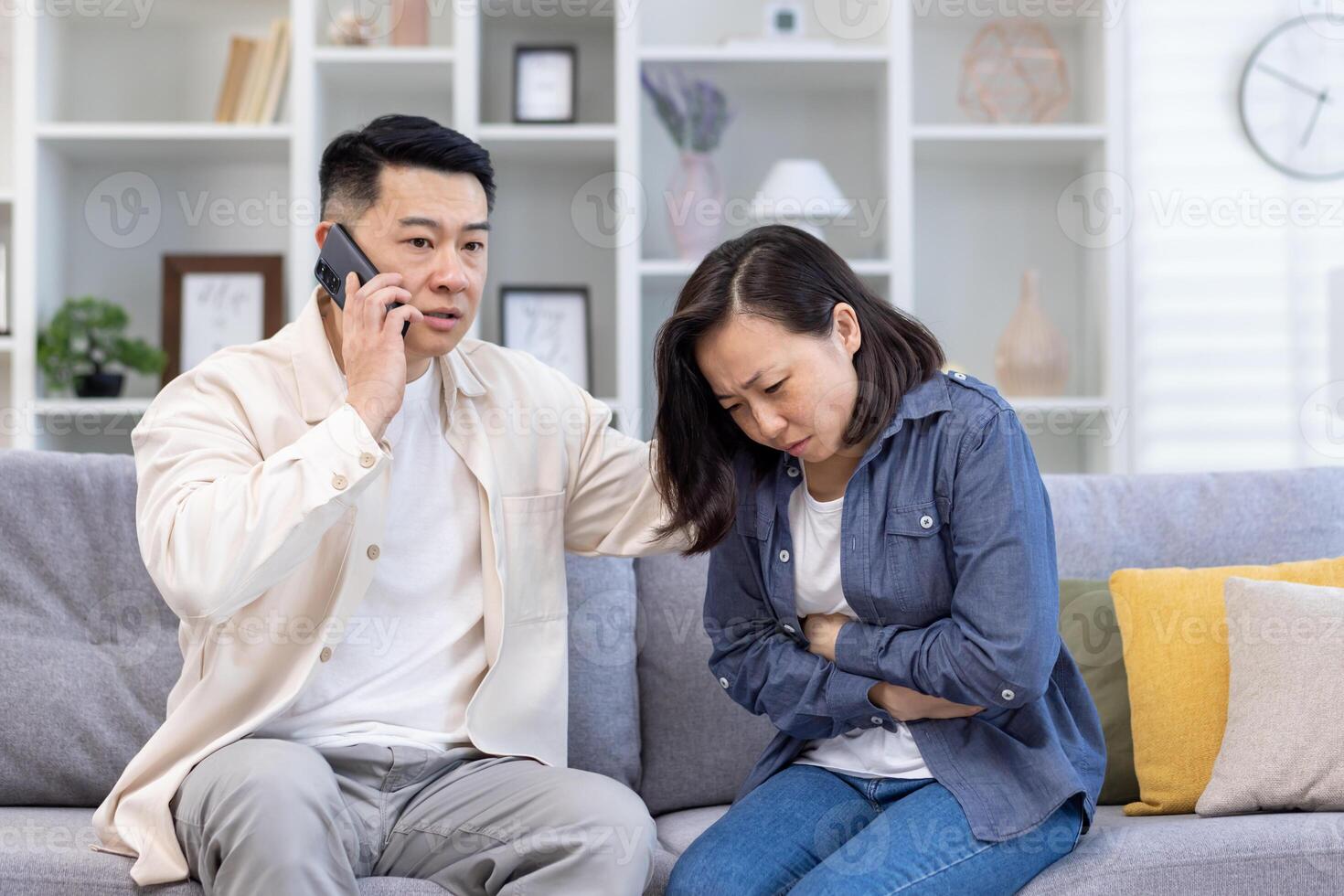 Young Asian family sitting on sofa at home. A woman holds her stomach, poisoning, pain, menstruation, pregnancy. The man calls an ambulance, calls a doctor. emergency call. photo