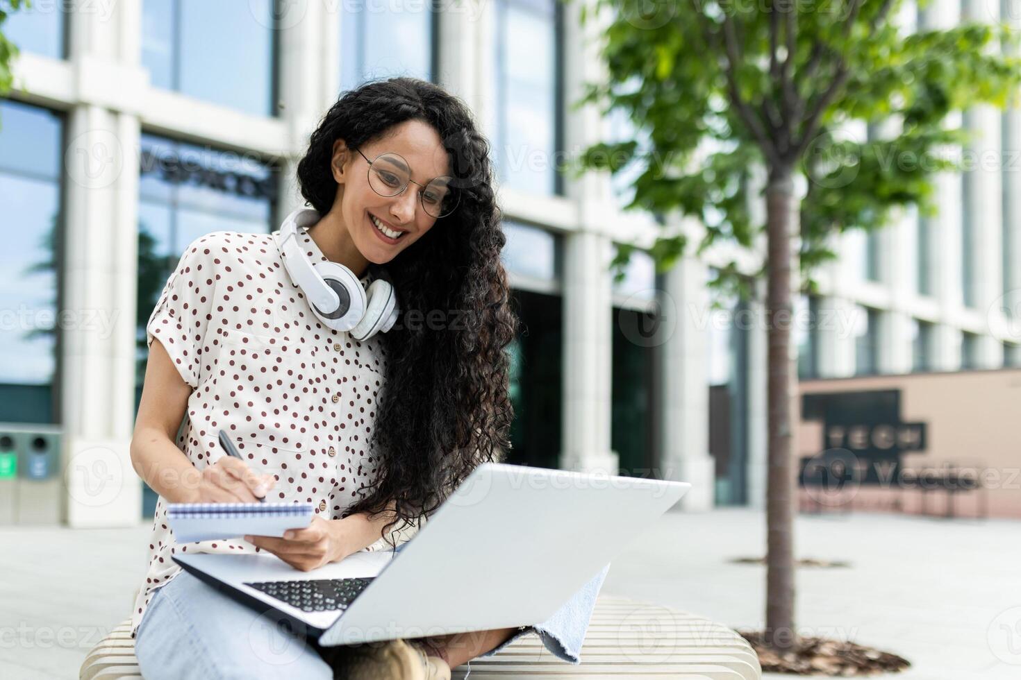 sonriente joven mujer con Rizado pelo utilizando ordenador portátil mientras sentado al aire libre en ciudad banco durante trabajo descanso. profesional y urbano estilo de vida. foto
