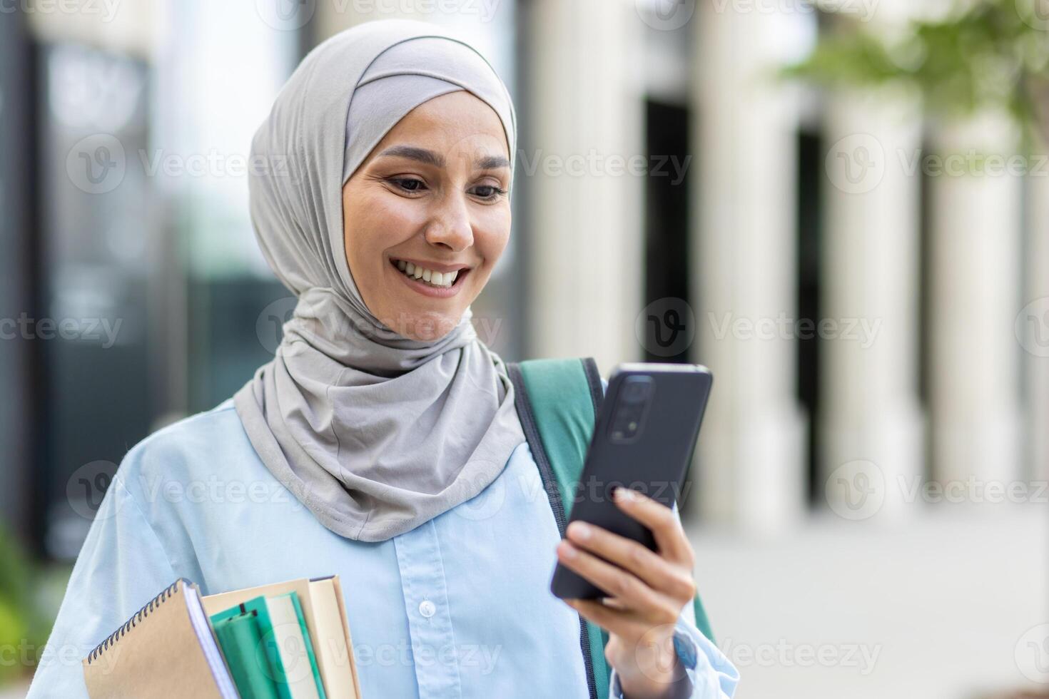 Young muslim woman in hijab walking outside university campus, female student smiling contentedly using app on phone, backpack on back and books in hands. photo