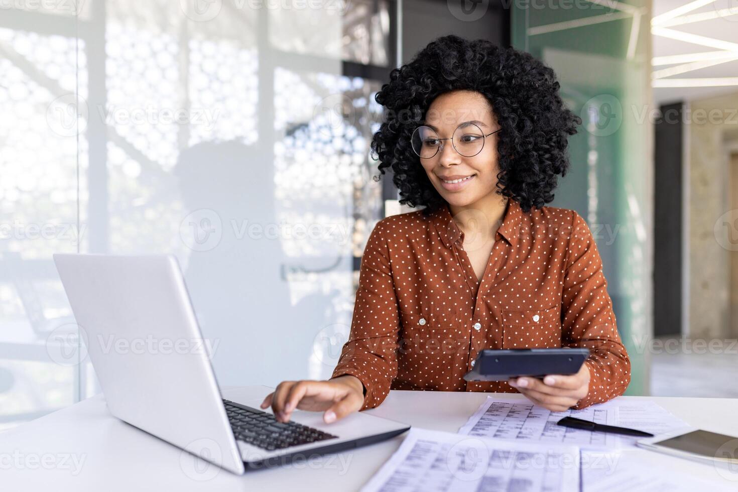 Young beautiful businesswoman, paper work inside office, female employee uses calculator at work, calculates and checks reports, accountant financier with laptop, reviews documents, uses laptop. photo