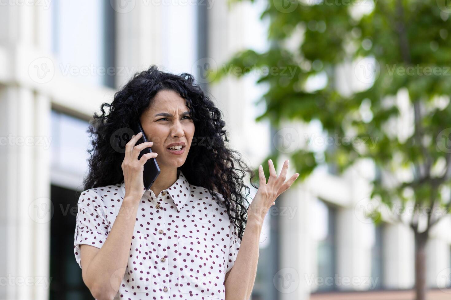 Frustrated woman in polka dot shirt outside making a confused gesture while talking on the phone, expressing misunderstanding. photo