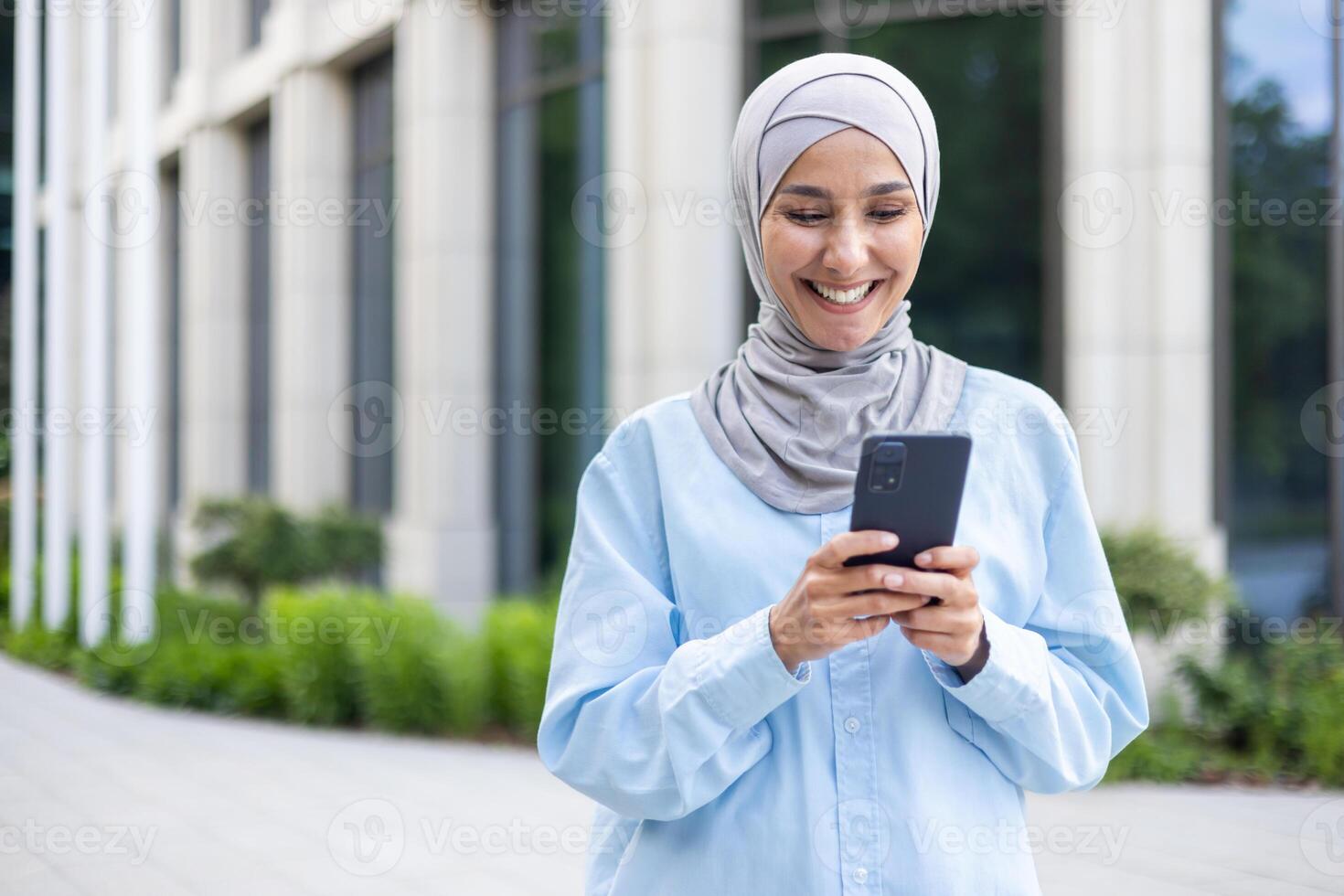 A young beautiful business woman in a hijab walks through the city, a Muslim woman holds a phone in her hands, uses an application on a smartphone, smiles, browses the Internet. photo