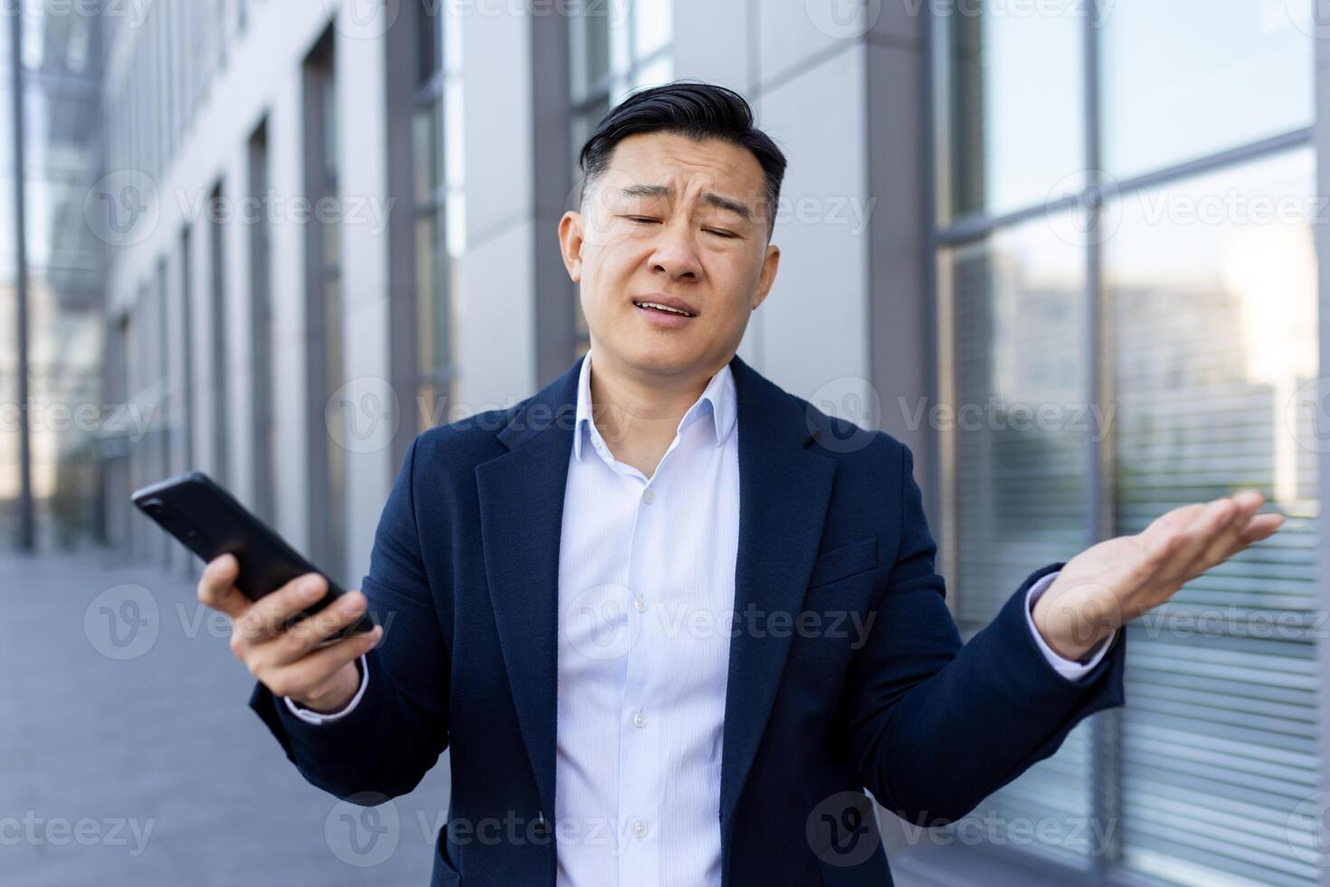Close-up photo of an upset young Asian male businessman standing in a suit outside an office building, holding a phone and waving his hands in frustration.