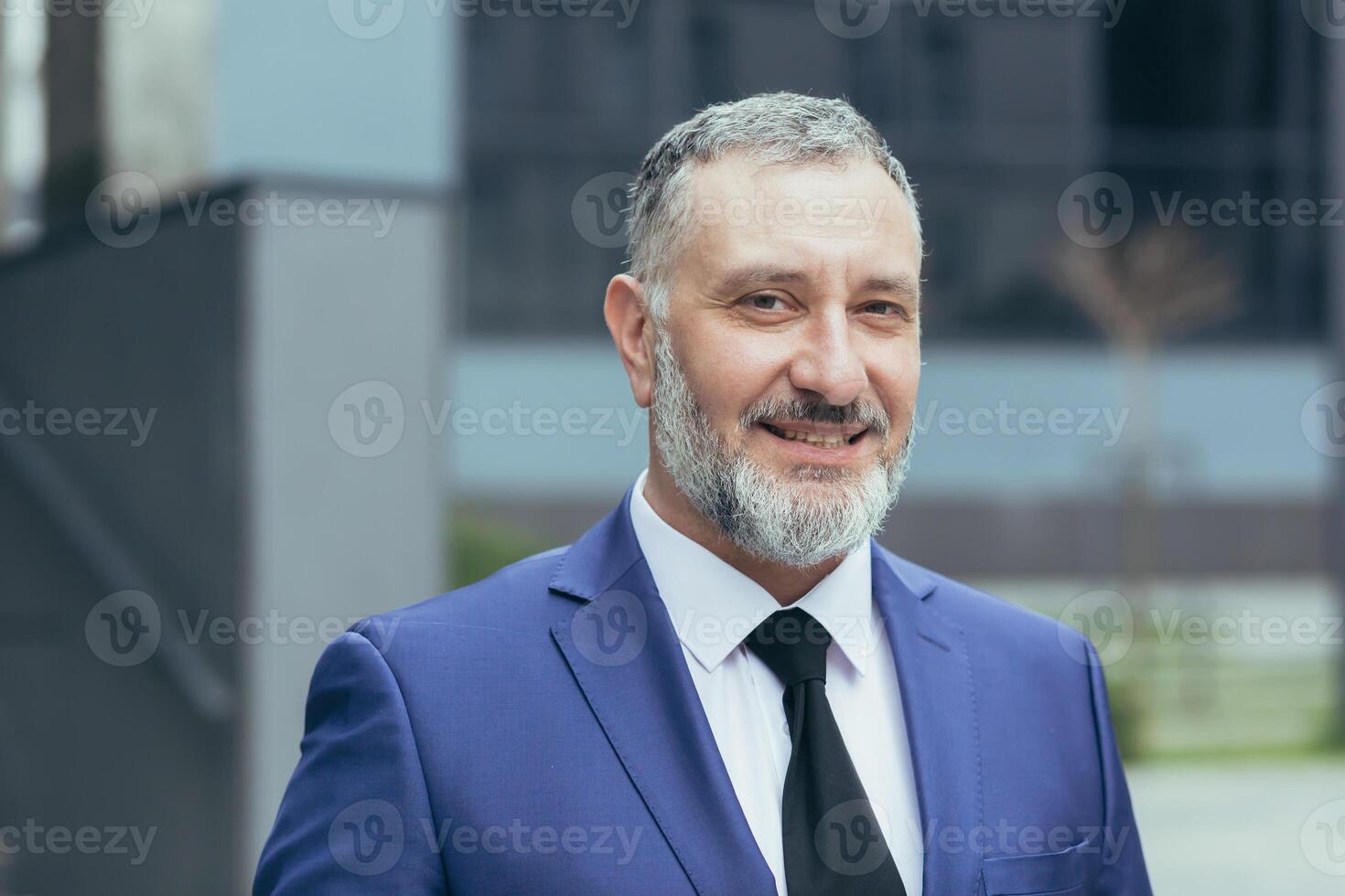 Portrait of senior handsome businessman man with gray hair and brown hair in business suit with tie. Near the office center, looks at the camera, smiles. photo