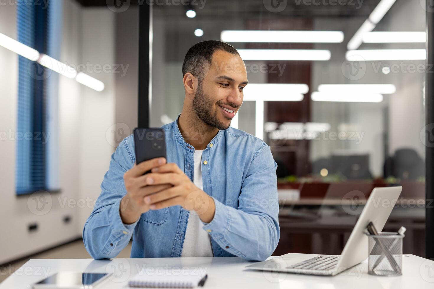 Smiling young arab man working in office on laptop, sitting at table and looking at screen, holding and using mobile phone. photo