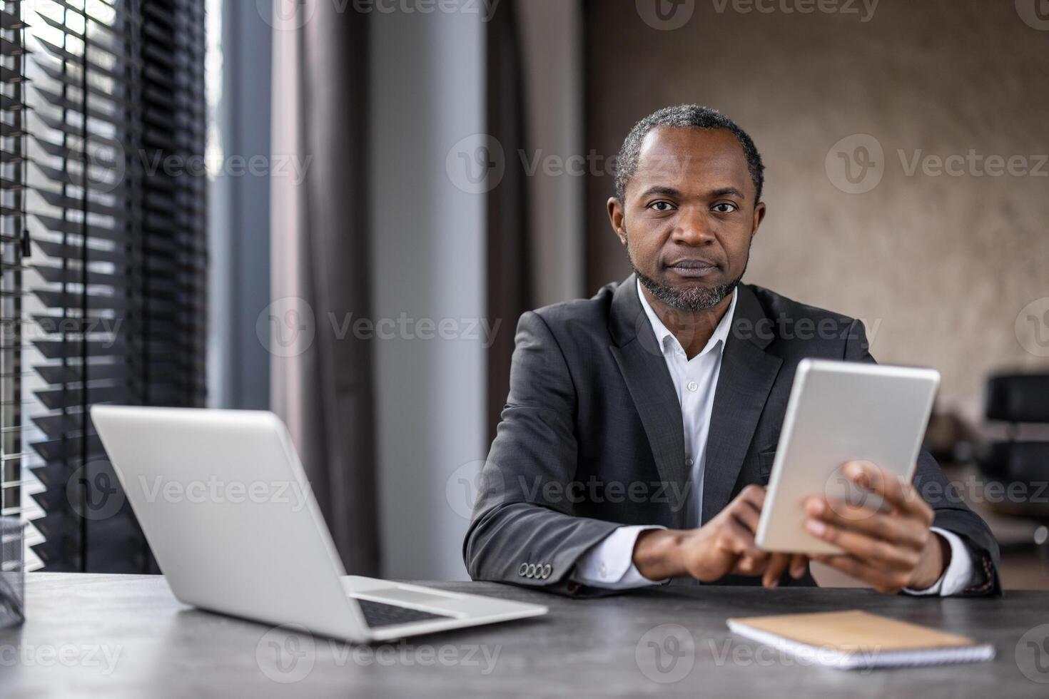 A man in a suit is sitting at a desk with two laptops and a tablet. He is focused on the tablet, possibly working on a project or browsing the internet. Concept of productivity and concentration photo