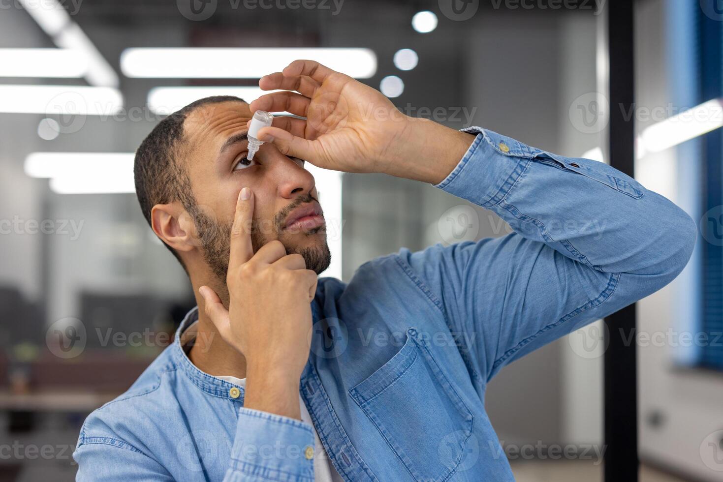 Close-up photo of a young Muslim man in a blue shirt sitting at a desk in a modern office and instilling medicine for inflammation and fatigue into his eye.