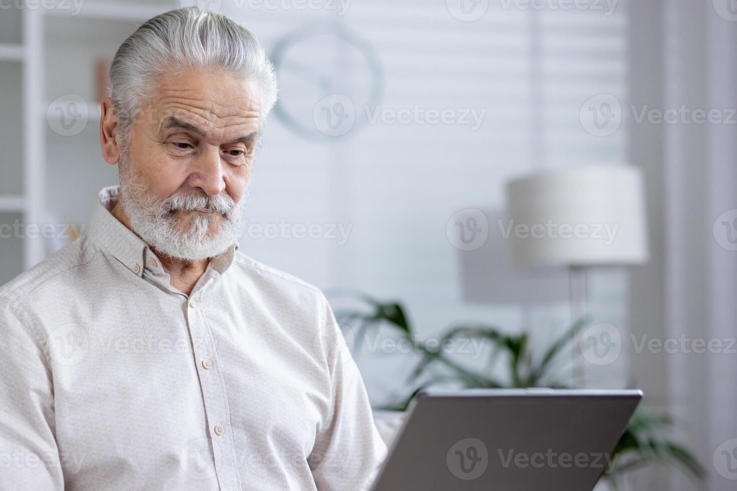 An elderly gentleman focuses intently on a digital tablet while seated in a well-lit, contemporary room, suggesting technology use among seniors. photo