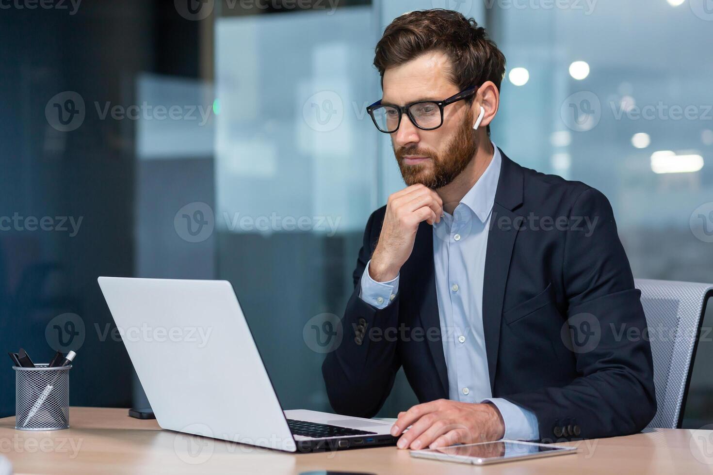 Senior businessman in glasses and beard is thinking about a decision, man is working inside the office and using laptop at work, mature investor in a business suit is sitting at the table photo