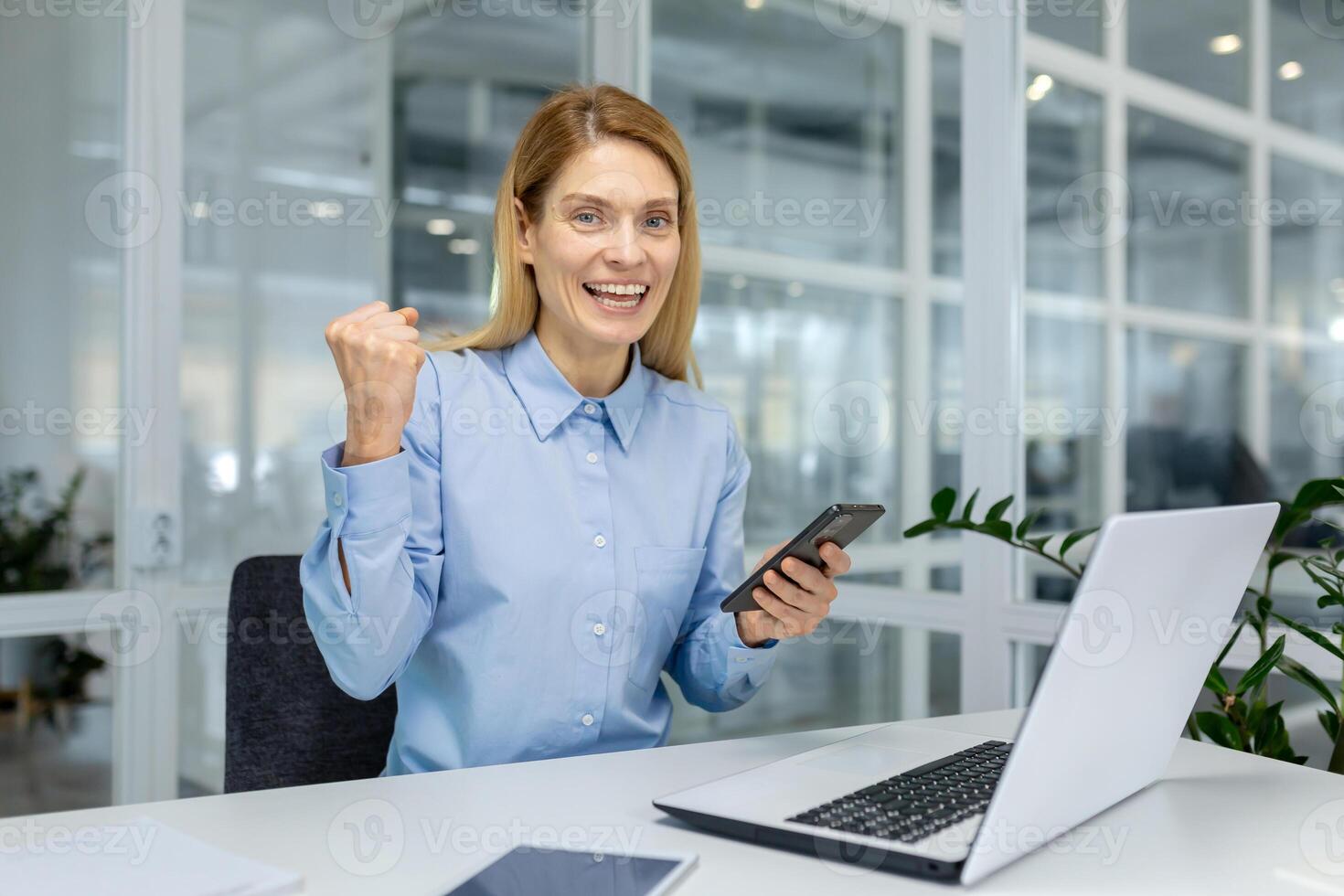 Positive lady in shirt making triumph gesture with hand while holding smartphone in modern workplace. Cheerful manager receiving pleasant text message with good news about job opportunities. photo