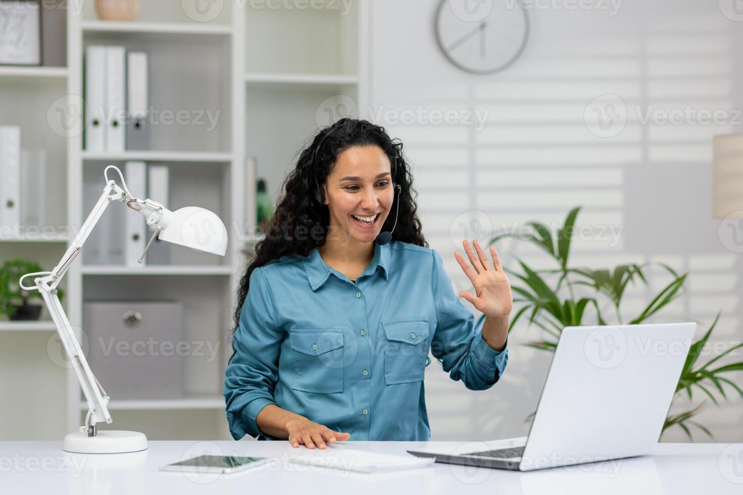 Friendly businesswoman in a smart blue shirt, waving and smiling during a virtual meeting in a well-lit office setting, showcasing effective remote work communication. photo