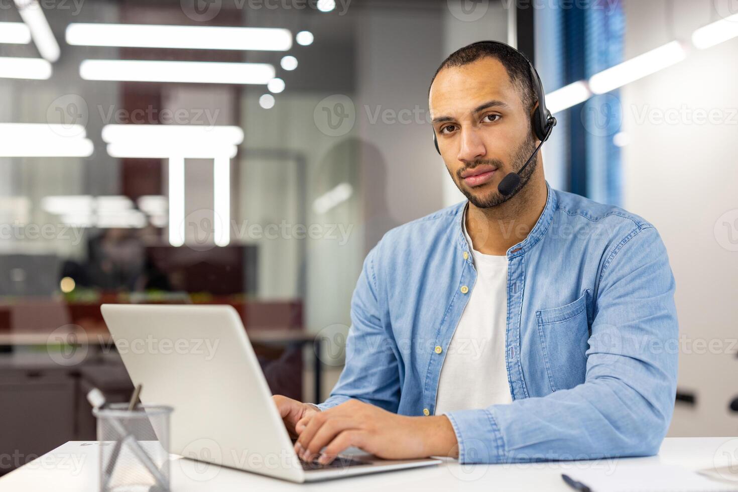 Portrait of a serious hispanic man in a shirt sitting in the office, at the table in front of the laptop and confidently looking at the camera.. photo