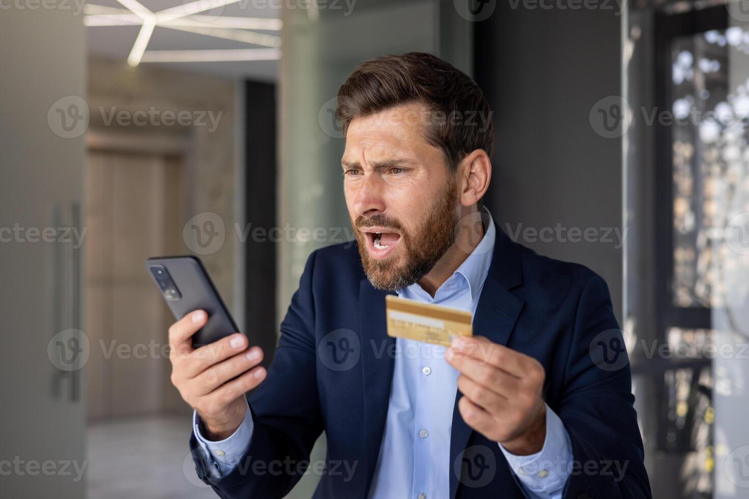 Close-up photo of angry young businessman in suit standing in office, holding golden credit card in hand and looking disappointedly at mobile phone screen.